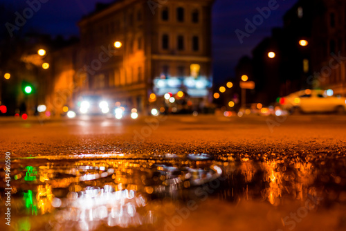 Nights lights of the big city, the night intersection between the houses on which cars travel. Close up view of a puddle on the level of the hatch