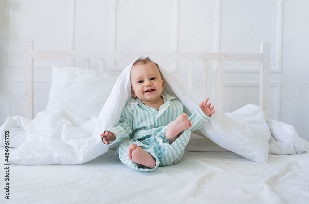 a cheerful little boy in striped pajamas is sitting and playing on a white bed in the room. Happy childhood a cheerful little boy in striped pajamas is sitting and playing on a white bed in the room. Happy childhood
