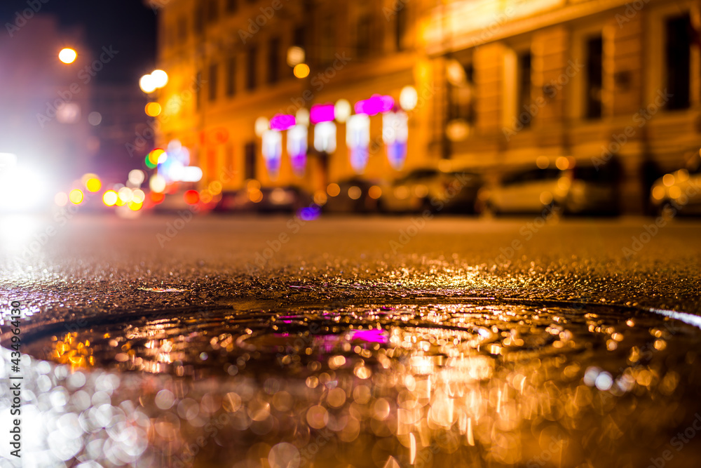 Nights lights of the big city, the city street with sparkling storefronts. Close up view of a puddle on the level of the hatch