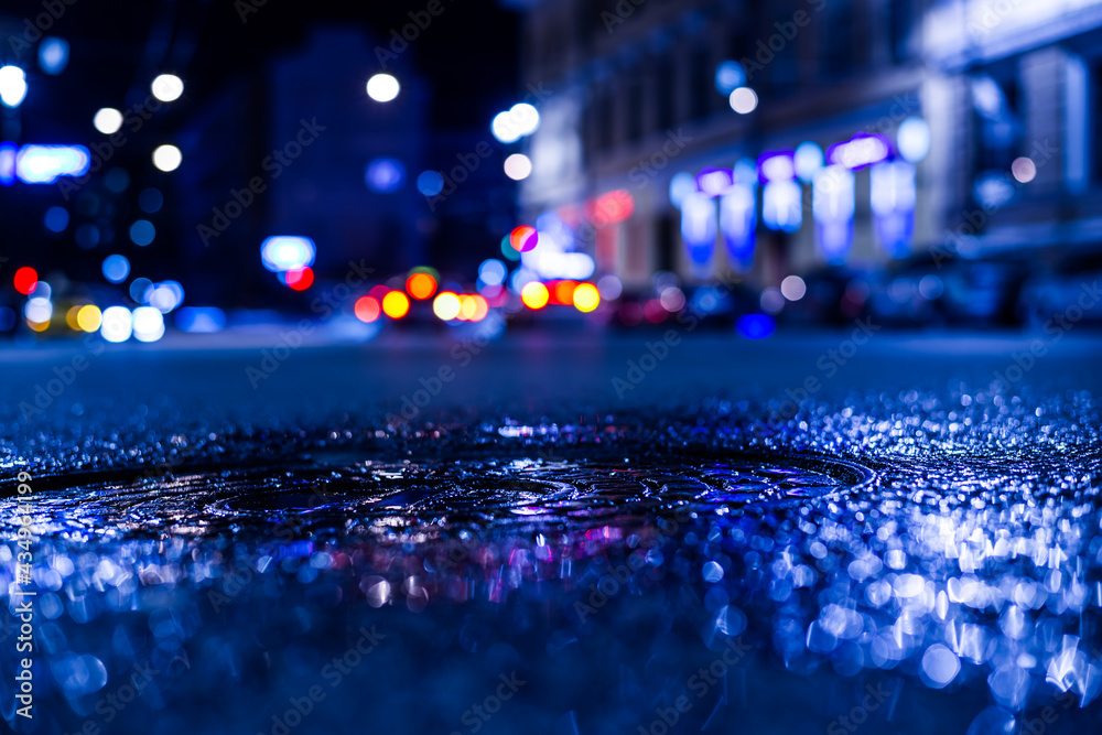Nights lights of the big city, the city street with sparkling storefronts. Close up view of a puddle on the level of the hatch