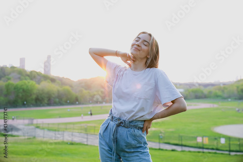 Happy young lady wearing jeans and a white t-shirt looking up and enjoying the weather. Sunlit photo with beautiful sunset in the background. Summer happiness. Green park in the background. © Anastasiia