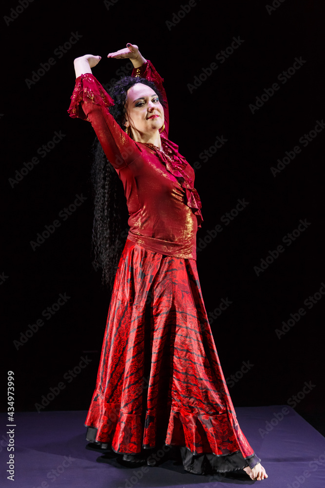 Gypsy woman with long black hair dances with her hands in motion in a red dress on a black background.