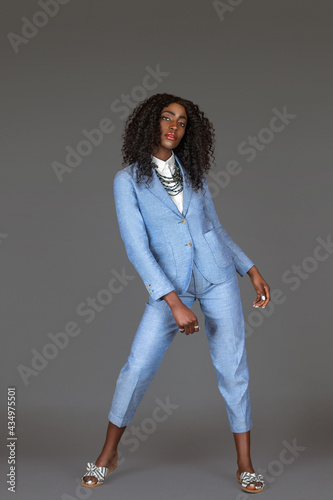Portrait of a tall sensual black woman with long curly black hair and beautiful makeup posing by herself in a studio with gray background wearing a blue suit with striped sandals and jewelry.