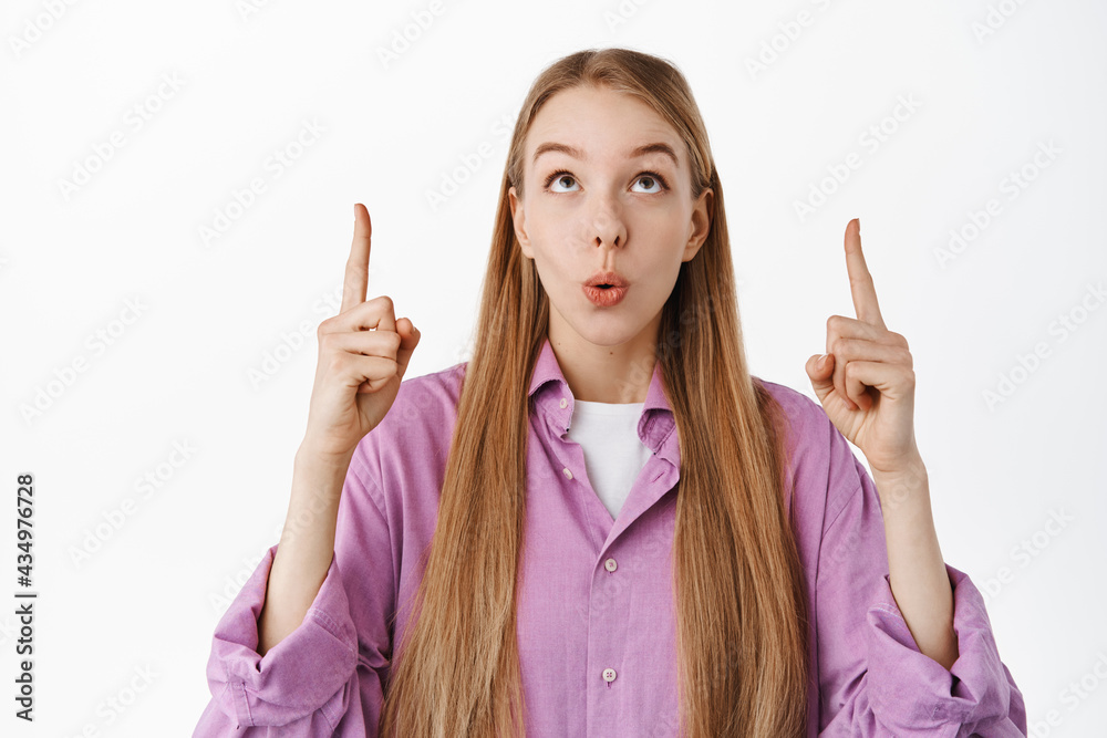 Close up of intrigued girl customer looking, pointing fingers up and say wow, checking out awesome special deal, advertisement, standing over white background Close up of intrigued girl customer looking, pointing fingers up and say wow, checking out awesome special deal, advertisement, standing over white background