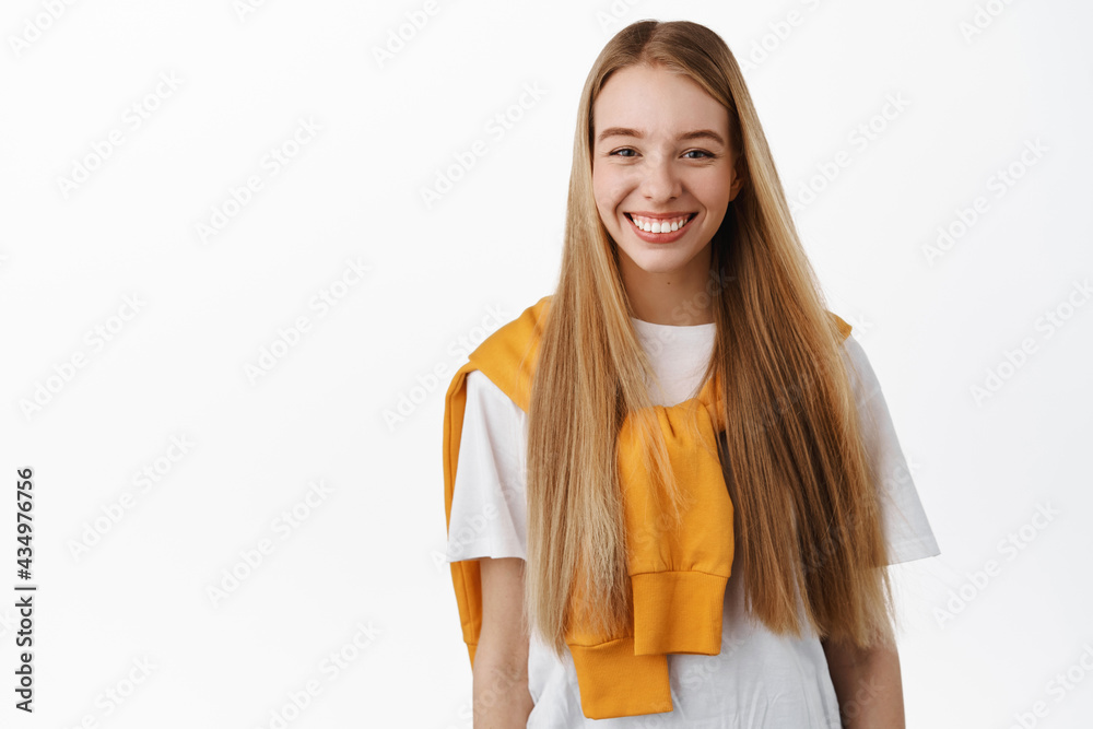 Beautiful young woman with natural blond long hair flying in air, girl looks at camera happy, laughing and smiling, standing against white background Beautiful young woman with natural blond long hair flying in air, girl looks at camera happy, laughing and smiling, standing against white background