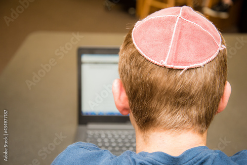 Young man wearing a yarmulke from the back doing work on a laptop in a library with colorful books on shelves. photo