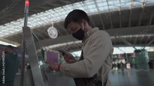 Man in face mask boarding on flight in airport. Traveler typing on touch screen of self check-in station in terminal. Flight registration, coronavirus outbreak. Self service machine at departure area photo