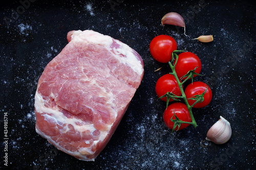 Raw piece of pork with red tomatoes, fresh garlic and salt on black background. Overhead view of Piece of fresh boneless pork, neck part or collar. Big piece of red raw meat on a black background.