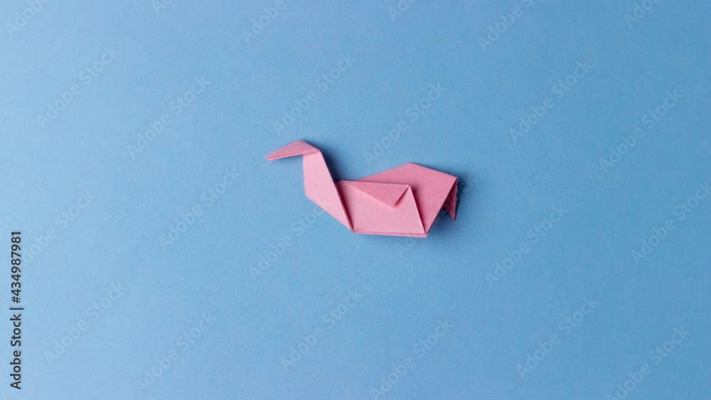The figure of the bird from a pink sheet in the origami style on a blue background, folded and and flies away. Step-by-step instruction. In center of the blue background. Pastime, hobby, activities