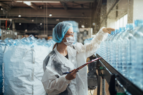 Female worker with protective face mask working in medical supplies research and production factory and checking canisters of distilled water before shipment. Inspection quality control.