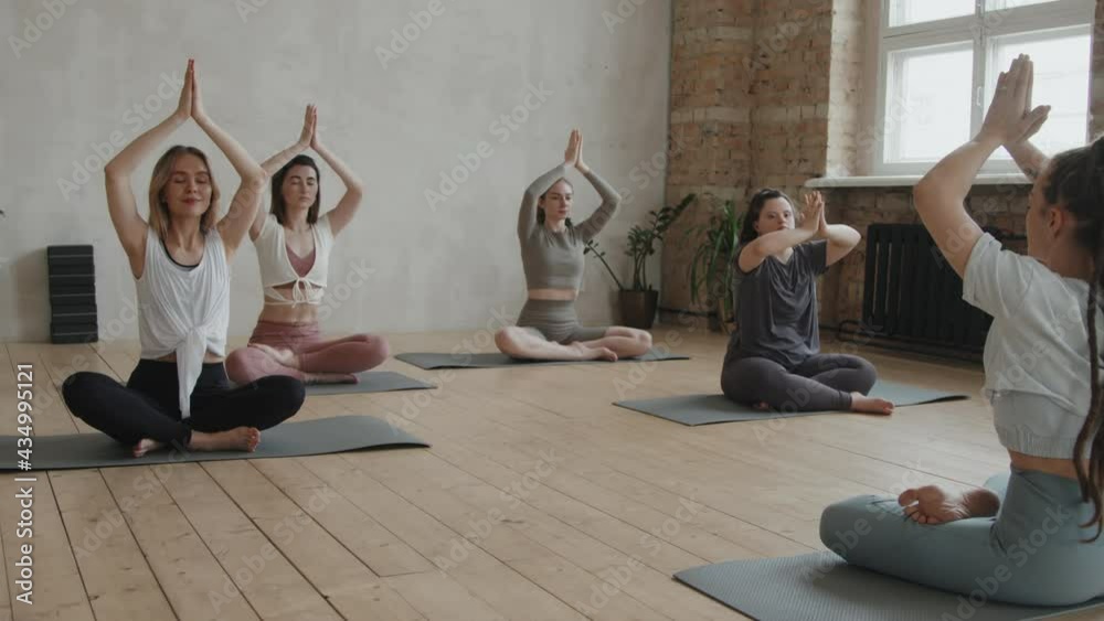Zoom-in slowmo of young woman with down syndrome practicing yoga in company of healthy girls and female coach sitting together on yoga mats in bright studio