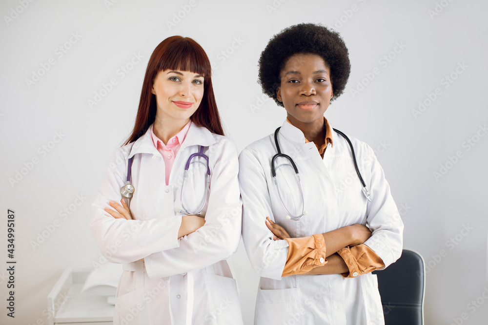 Two smiling happy African and Caucasian female doctors colleagues, wearing white coats, standing side by side with folded arms, posing at camera at modern light medical office Two smiling happy African and Caucasian female doctors colleagues, wearing white coats, standing side by side with folded arms, posing at camera at modern light medical office