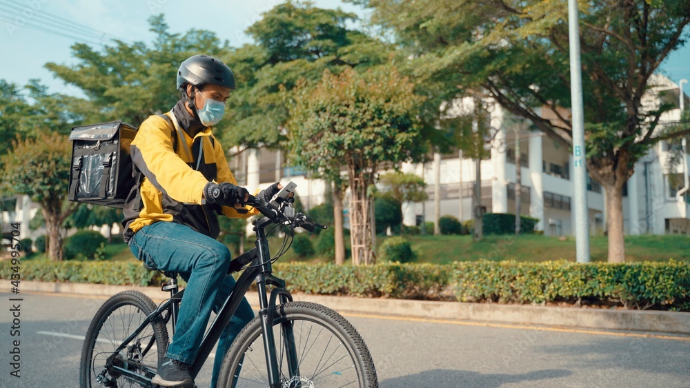 Obraz premium Happy Food Delivery Courier Wearing Thermal Backpack Rides a Bike on the Road To Deliver Orders for Clients and Customers. Sunny Day in Modern City with Stylish Urban Buildings.