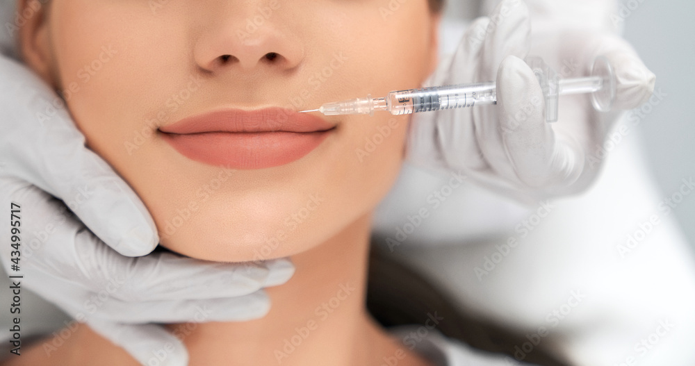 Close up of beautician in white rubber gloves doing injection in lips for young beautiful woman. Concept of process augmentation and improvement lips in professional salon. 