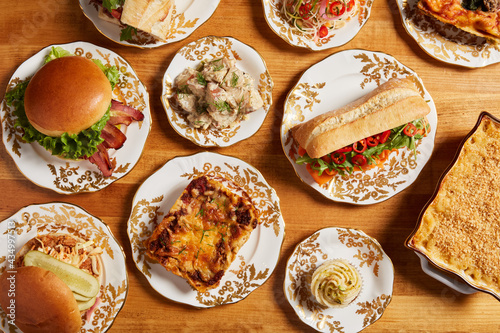 Top view of an assortment of food including a smoked carrot sandwich, lasagna, BLT, mac n cheese, cupcake and other savory items served on white plates placed on a wooden surface