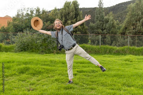 young traveler with glasses posing amused smiling carrying his camera and holding his hat with his arms outstretched full-length.