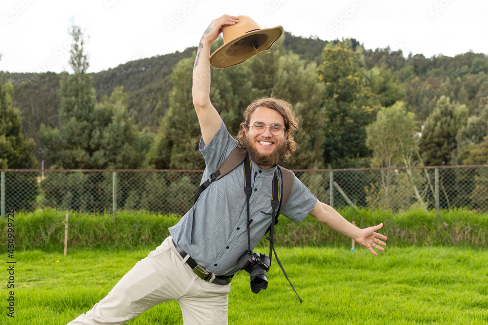 young male traveler with glasses posing amused smiling carrying his camera and holding his hat