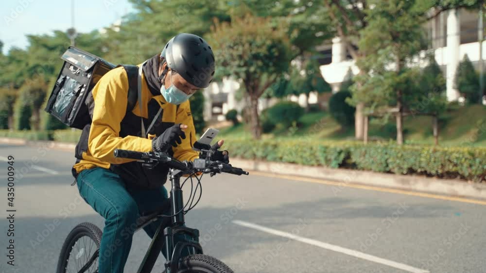 Asian delivery man in mask searching address with mobile phone on city street. Bike courier using smartphone for delivering food outdoors. Uniform guy looking around in urban background. Asian delivery man in mask searching address with mobile phone on city street. Bike courier using smartphone for delivering food outdoors. Uniform guy looking around in urban background.