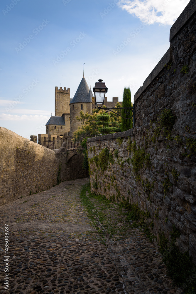 Medieval castle towers stand out in the background over the stone walls of an cobbled alley in the Cite of the fortified city of Carcassonne at sunrise, UNESCO World Heritage Site, France