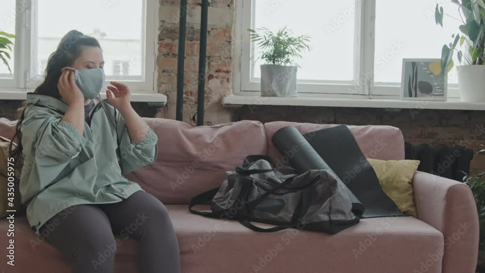Medium shot of young woman with down syndrome putting on face mask, taking her sports bag and leaving modern living room Medium shot of young woman with down syndrome putting on face mask, taking her sports bag and leaving modern living room