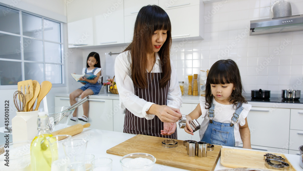 Asain lovely family, mom and two little daughters cooking and playing in modern kitchen. The younger sister learning how to make a bakery or bread while the elder sitting on counter and reading book Asain lovely family, mom and two little daughters cooking and playing in modern kitchen. The younger sister learning how to make a bakery or bread while the elder sitting on counter and reading book