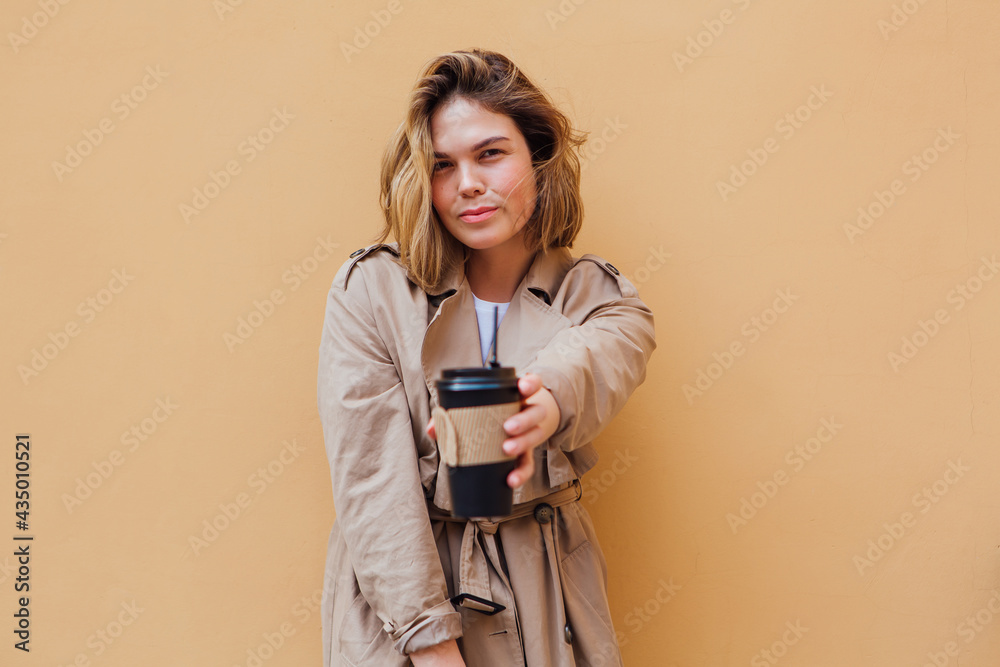 Young millennial woman with wild hair dressed in an autumn coat standing a cup of coffee to go near the wall. Young millennial woman with wild hair dressed in an autumn coat standing a cup of coffee to go near the wall.