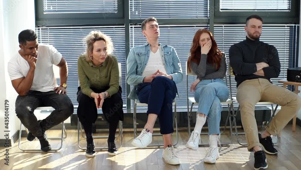 Front view of group of stressed five young diverse multiethnic men and women waiting job interview sitting in chairs in modern office lobby on background of window and bright sunlight. 