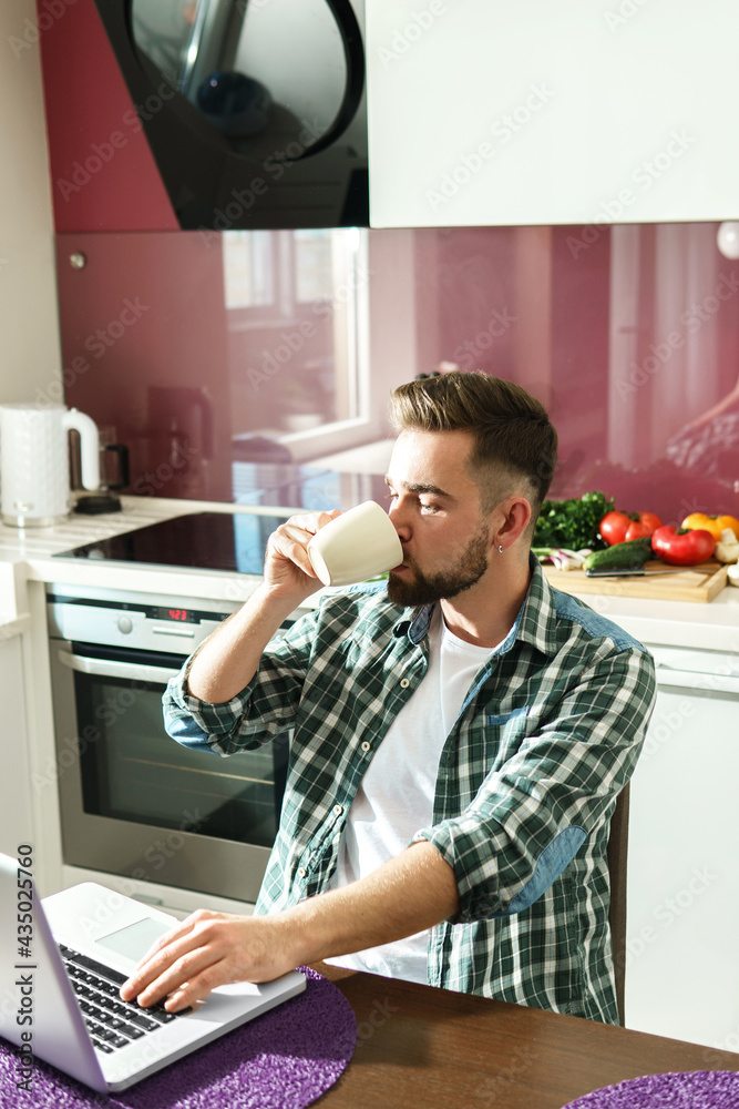 Handsome man drinking coffee or tea and using laptop in the kitchen