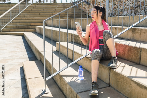 young caucasian sportswoman sitting on concrete steps and using her phone. © dzmatph