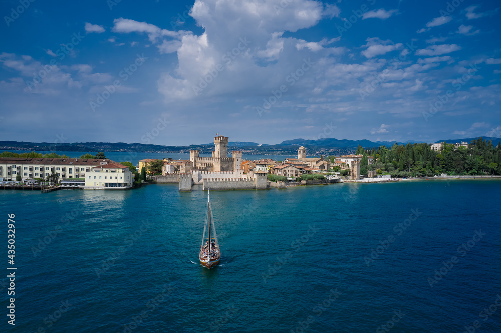 The Old Sailing Ship is sailing near Sirmione Castle. Rocca Scaligera Castle in Sirmione Lake Garda Italy. Clouds in the blue sky. Aerial view.