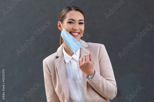 Beautiful businesswoman, young woman, in medical protective mask taking off from face, in formal jacket standing outdoors, looking at camera. Coronavirus, virus, epidemic, pandemic covid-19 concept photo