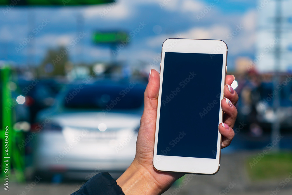 Girl holds a smartphone in his hands. Mock up phone with white screen against the background of an electric car at a charging station. Girl holds a smartphone in his hands. Mock up phone with white screen against the background of an electric car at a charging station.