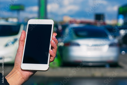 Girl holds a smartphone in his hands. Mock up phone with white screen against the background of an electric car at a charging station.