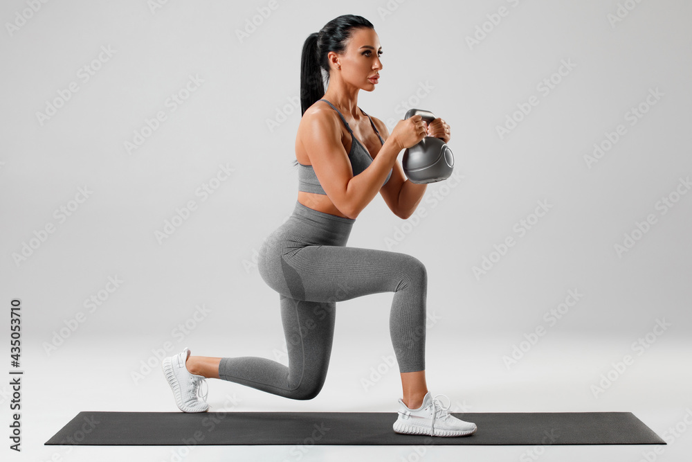 Fototapeta premium Active girl doing lunges exercises with kettlebell, leg muscle training. Fitness woman doing front forward one leg step lunge