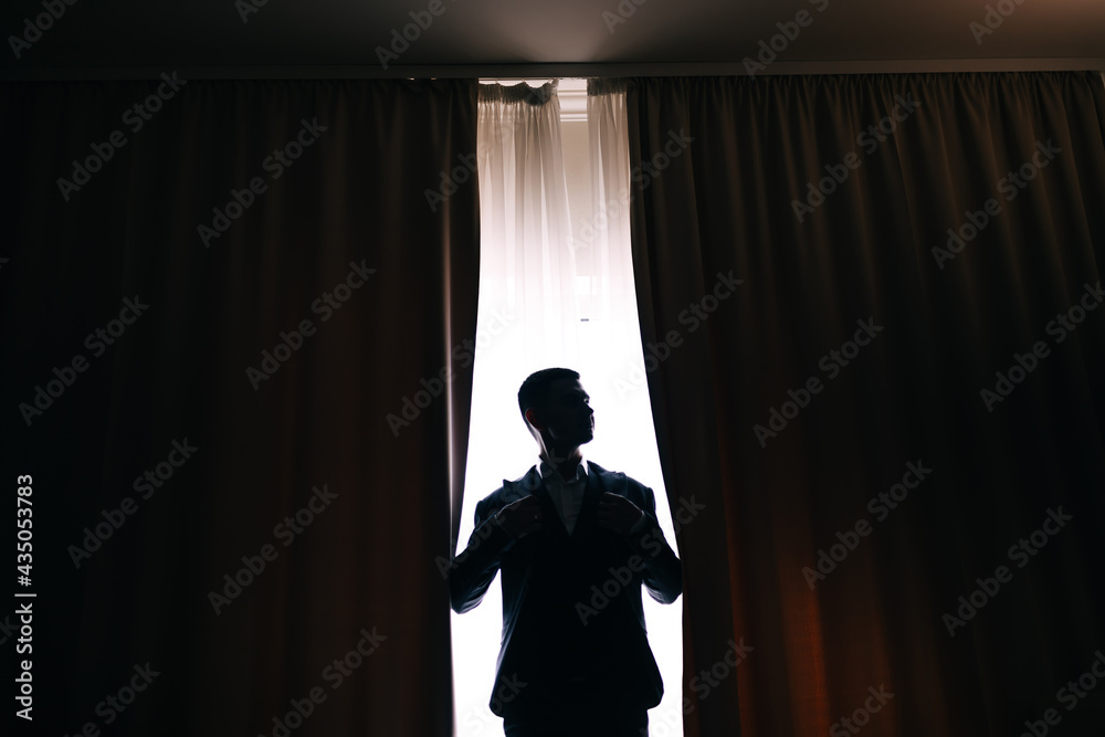 Stylish groom silhouette in suit posing at window light. Confident and happy portrait of man. Groom getting ready in morning