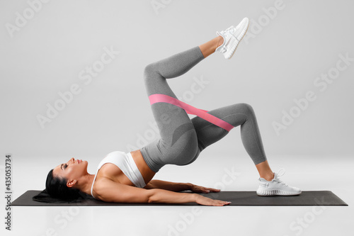 Athletic girl doing glute bridge exercise with resistance band on gray background. Fitness woman working out photo