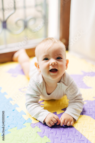 Surprised joyful baby lies on his tummy on a colored rug on the floor against the background of a window