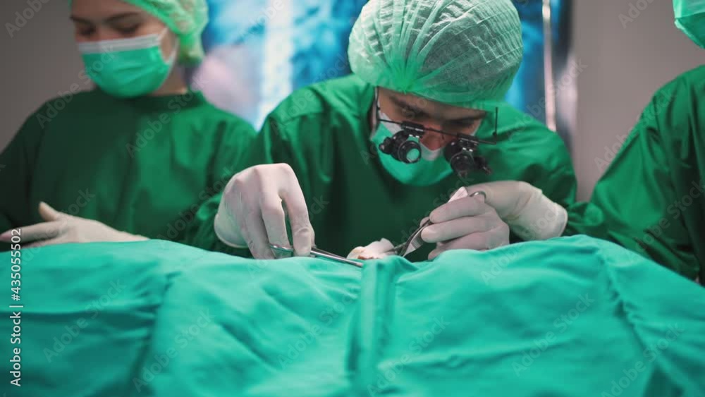 Group of professional doctor wearing uniform and face mask doing surgery in the hospital in the operating room under light. Doctor using scisor and forceps to help patient. Medical health and cure Group of professional doctor wearing uniform and face mask doing surgery in the hospital in the operating room under light. Doctor using scisor and forceps to help patient. Medical health and cure