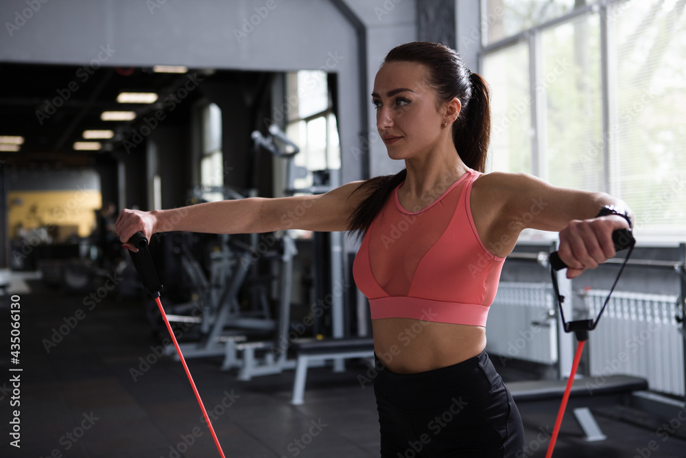 Fototapeta premium Female bodybuilder warming up shoulders with resistance band before working out at gym, copy space
