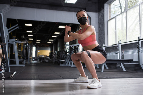 Sportswoman wearing medical face mask, doing squats with resistance band, working out at gym during coronavirus pandemic