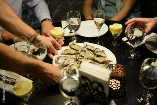 Hands putting plate with many oysters on table. People sitting by table,  with glasses and candle. © Oskars