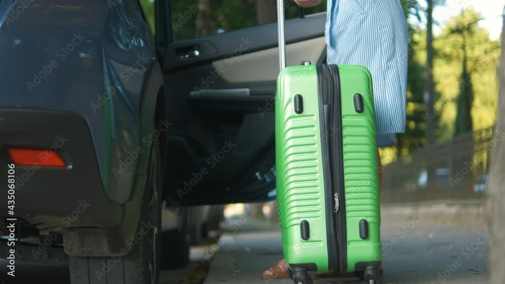Young woman driver putting green suitcase inside car on city street. Travel and vacations concept.