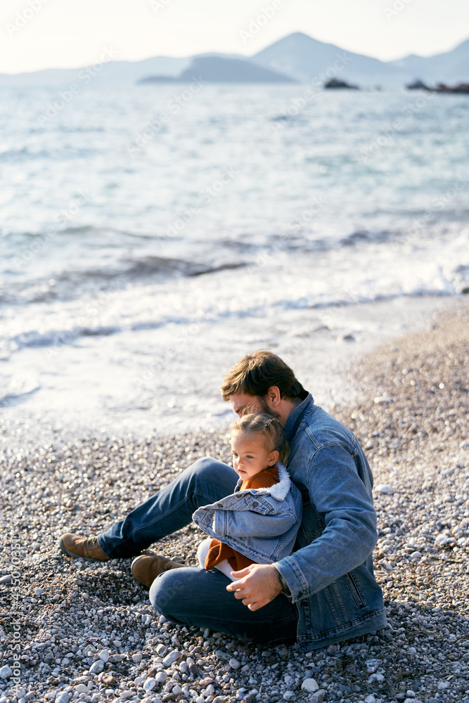 Dad in a denim jacket sits on a pebble beach and holds a little girl in his lap. Side view