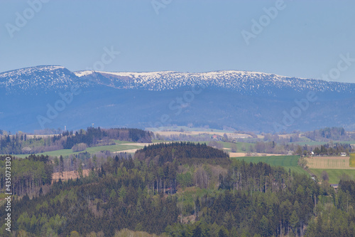 Landscape. Green forest. Hills. Fields. Snow-capped mountain peaks. Small houses. Blue sky.