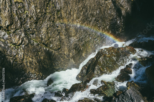 Atmospheric landscape with rainbow above turbulent mountain river among rocks with moss near rocky wall in sunshine. Beautiful alpine scenery with mossy stones in powerful mountain river in sunlight.