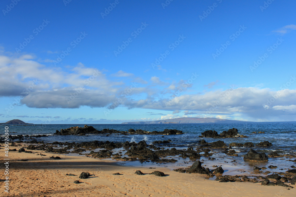 Sandy beach with black volcanic rocks in the Pacific ocean water on Maui Island, Hawaii and view to the islands Moloka’i and Lanai in the blurred background Sandy beach with black volcanic rocks in the Pacific ocean water on Maui Island, Hawaii and view to the islands Moloka’i and Lanai in the blurred background