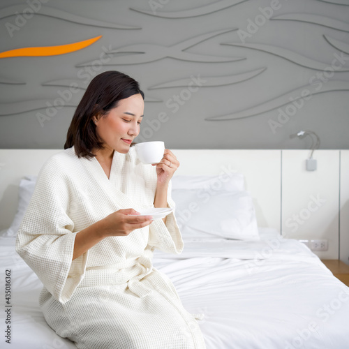 Asian woman in a bathrobe drinking hot coffee on bed in the morning.