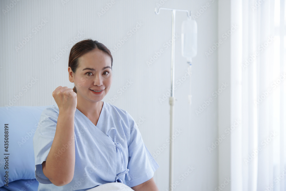 Portrait of Asian young patient woman smiling and raising a fist means fighting. She is sitting on bed with saline bag on stand in hospital room. Portrait of Asian young patient woman smiling and raising a fist means fighting. She is sitting on bed with saline bag on stand in hospital room.