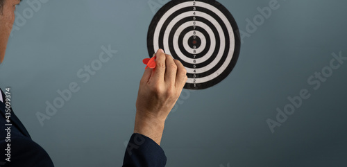 In the right hand of a man in a suit, there is a red dart that is being pinned into the center of a circular target on the wall, The goal of concep is to achieve it.