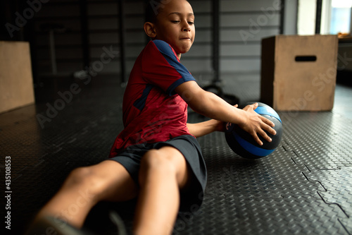 Focused motivated strong black little boy working out in gym, having determined look, training abs, doing crunches holding medicine ball. Dark skinned kid using fitness equipment for core muscles photo